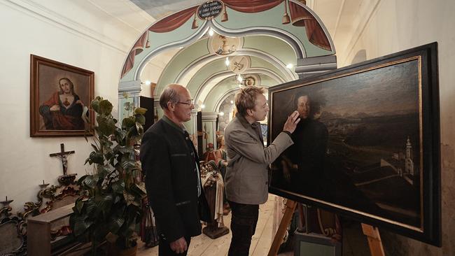 Max Müller mit Pfarrgemeinderat Josef Urschitz vor einem Bild, auf dem der Goldpfarrer von Latschach zu sehen ist.