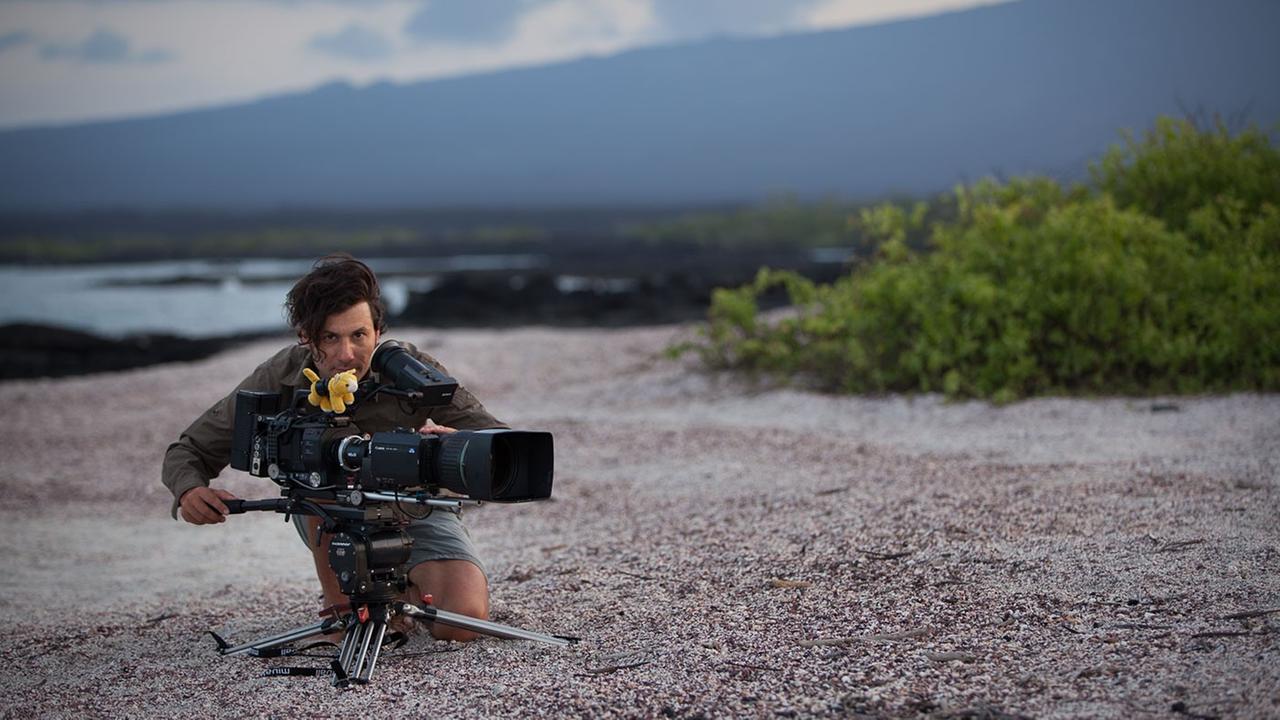 Der Kameramann Michael Riegler an seiner Kamera sitzend. Im Hintergrund eine Küstenlandschaft.