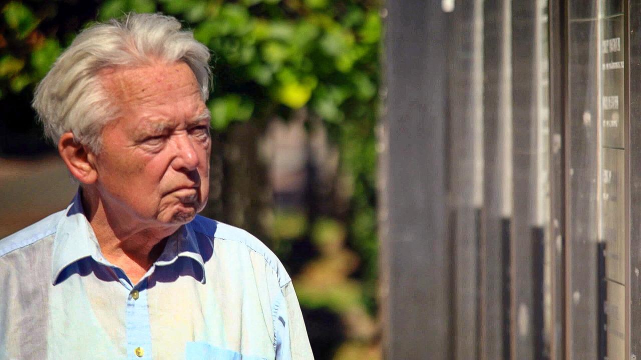 © ORF Hans Haider vom Verein Erinnern Villach steht im Freien neben einer Wand mit Gedenktafeln