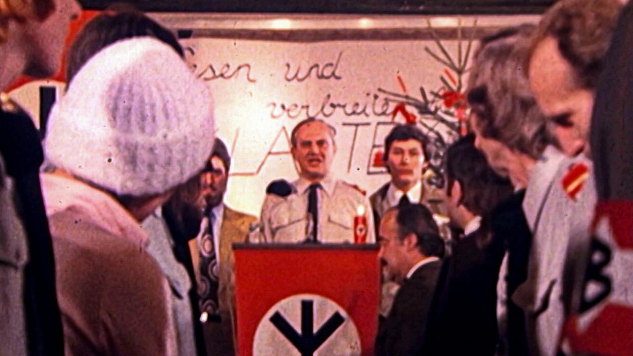 © ORF Veranstaltung der NDP 1977 mit Menschen in Uniform um einen Rednerpult mit Symbol und Schriftzug im Hintergrund.