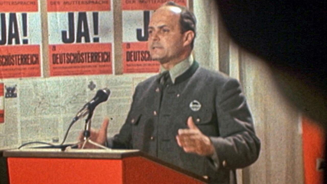 © ORF NDP-Gründer Norbert Burger 1977 spricht am Rednerpult vor Plakaten mit der Aufschrift „JA!“.