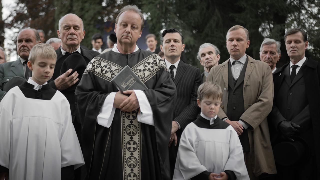Ein Priester hält bei einer Trauerfeier ein Buch, umgeben von Ministranten und schwarz gekleideten Trauergästen.