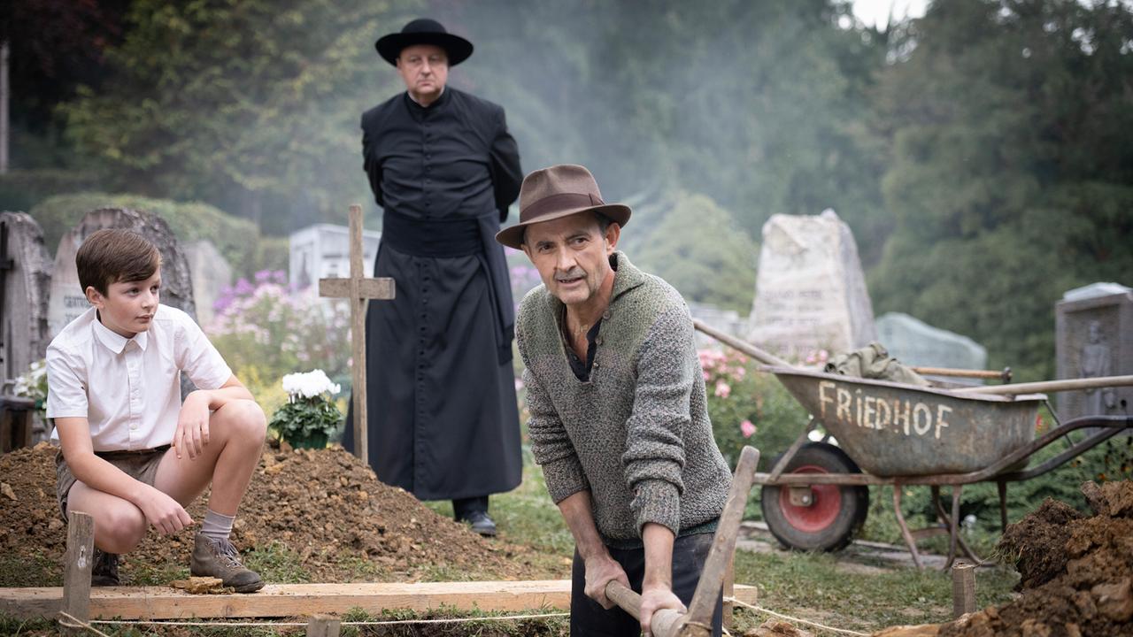 Josef gräbt ein Grab, während der Junge Michael und Hochwürden daneben auf einem Friedhof stehen.