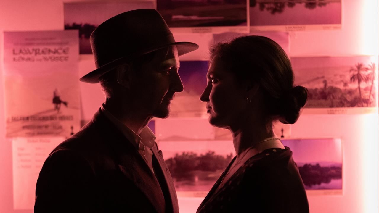 Josef und Ragusa stehen sich in rotem Licht sehr nah gegenüber, im Hintergrund sind Fotografien an einer Wand aufgeklebt.