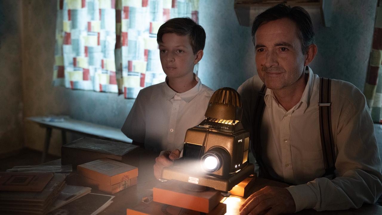 Michael und Josef sitzen nebeneinander in einem verdunkelten Raum an einem Tisch, auf dem ein Filmprojektor eingeschaltet ist und Licht abstrahlt.
