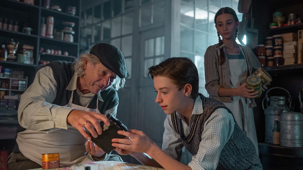 Beim Gemischtwarenhändler begutachtet der 12-jährige Josef eine Filmkamera, während die 13-jährige Ragusa im Hintergrund Konserven hält und ihm zusieht.