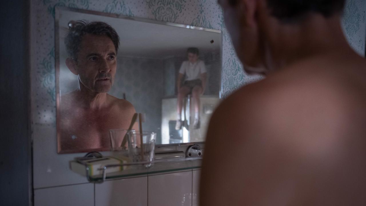 Josef steht mit nackten Oberkörper vor einem Badezimmerspiegel, in dem der im Hintergrund sitzenden Michael zu sehen ist.