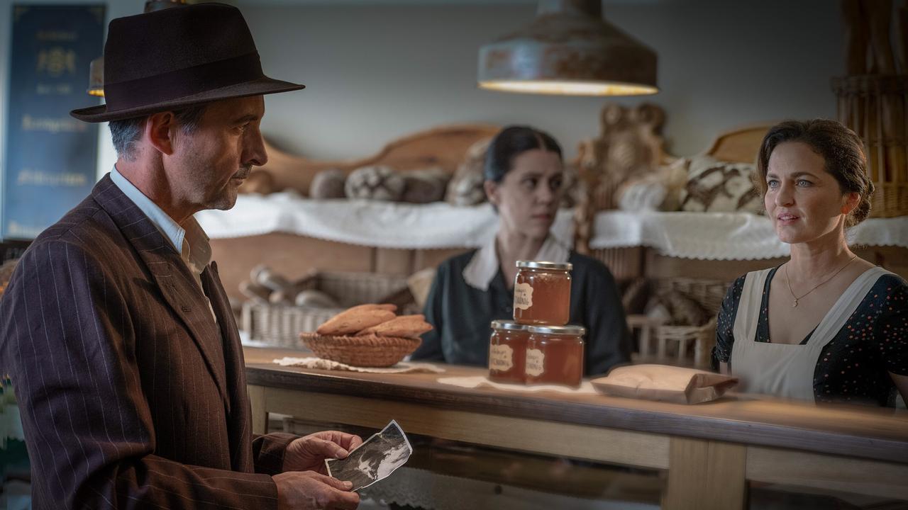 Josef mit Hut steht an einer Bäckertheke, hinter der Ragusa und eine weitere Frau Gebäck verkaufen.
