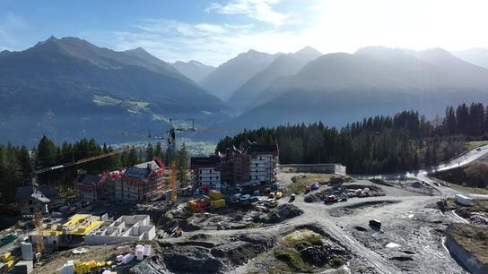 Die Großbaustelle des Luxusresorts Kitzbühel Alps am Pass Thurn liegt eingebettet in einer alpinen Landschaft, während im Hintergrund Berge und Wälder unter sonnigem Himmel sichtbar sind.