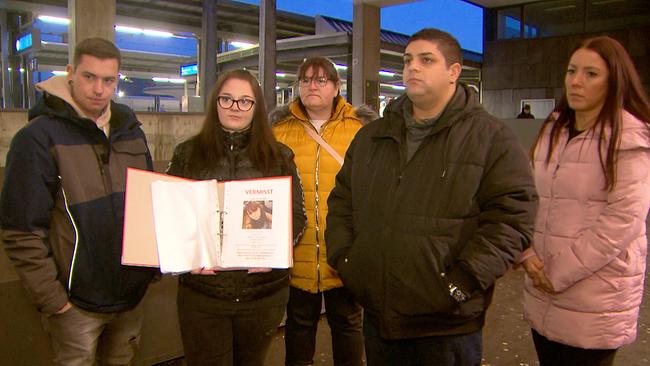 Acht Monate suchte die Familie vergeblich nach Christa: Fünf Personen stehen an einem Bahnhofsvorplatz in Winterjacken, eine hält einen Ordner mit einem „Vermisst“-Aushang und Foto hoch.