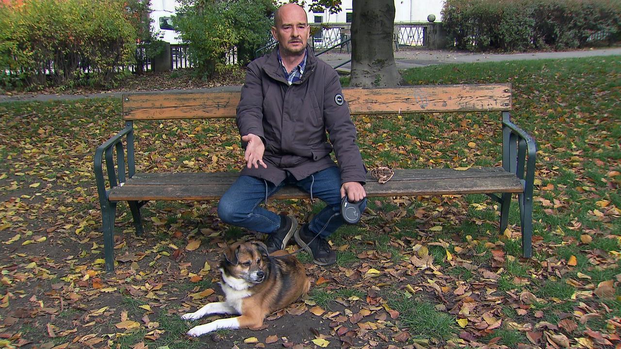 Auf einer Parkbank sitzt Herr Andreas in einer Jacke und Jeans, während sein Hund Benno an der Leine entspannt davor im herbstlichen Laub liegt.