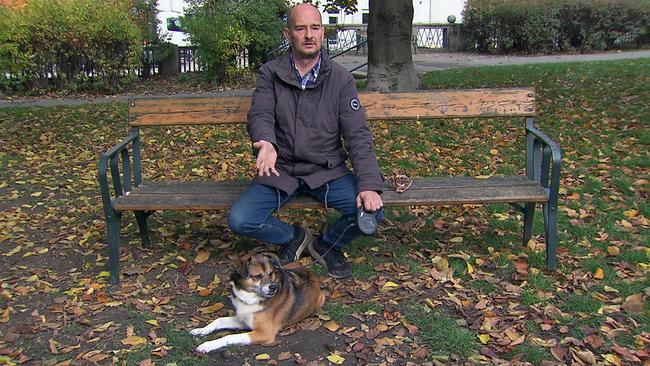 Auf einer Parkbank sitzt Herr Andreas in einer Jacke und Jeans, während sein Hund Benno an der Leine entspannt davor im herbstlichen Laub liegt.