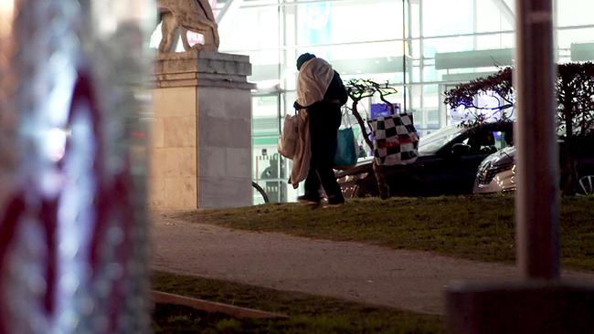 Ein junger Obdachloser geht nachts im Freien mit mehreren Plastiksackerln und Decken vor einem hell beleuchteten modernen Gebäude.