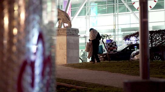 Ein junger Obdachloser geht nachts im Freien mit mehreren Plastiksackerln und Decken vor einem hell beleuchteten modernen Gebäude.