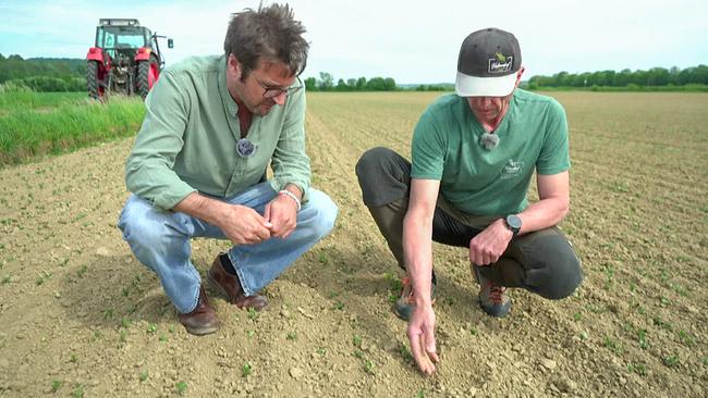 © ORF Landwirt Christian Gartner und ein Reporter hocken auf einem frisch gepflanzten Feld und begutachten die Erde, im Hintergrund steht ein Traktor.