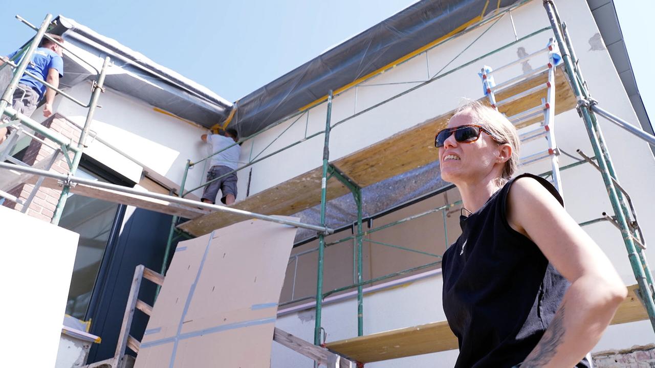 © ORF Nicole Bauer mit Sonnenbrille steht vor ihrem eingerüsteten Haus, an dessen Fassade Bauarbeiter arbeiten.