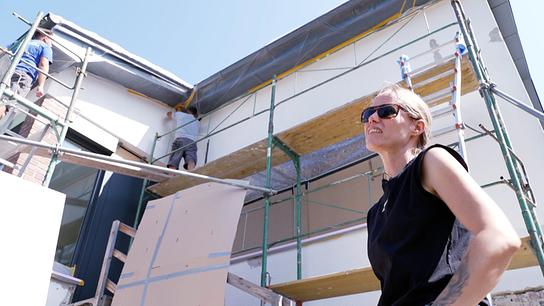 Nicole Bauer mit Sonnenbrille steht vor ihrem eingerüsteten Haus, an dessen Fassade Bauarbeiter arbeiten.