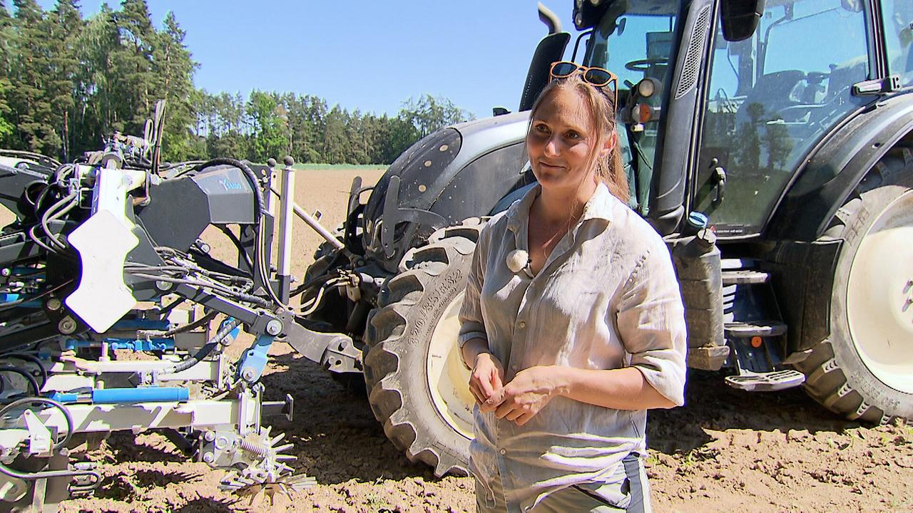 © ORF Viktoria Hutter steht bei Sonnenschein auf einem Feld vor einem großen Traktor.
