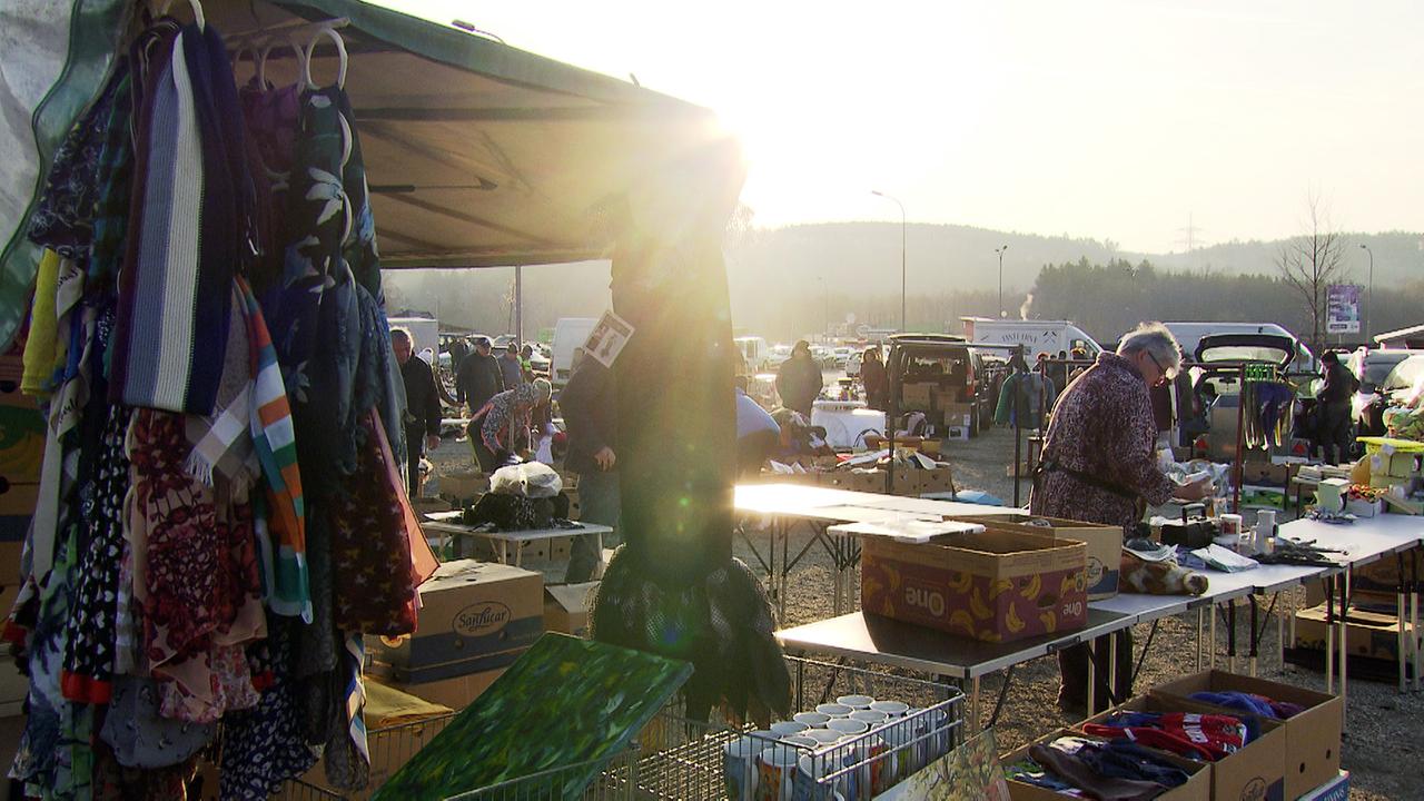 Zu sehen sind Flohmarktstandler, die im Morgengrauen ihre Tische auf einem großen Platz aufbauen und Ihre Waren auspacken.