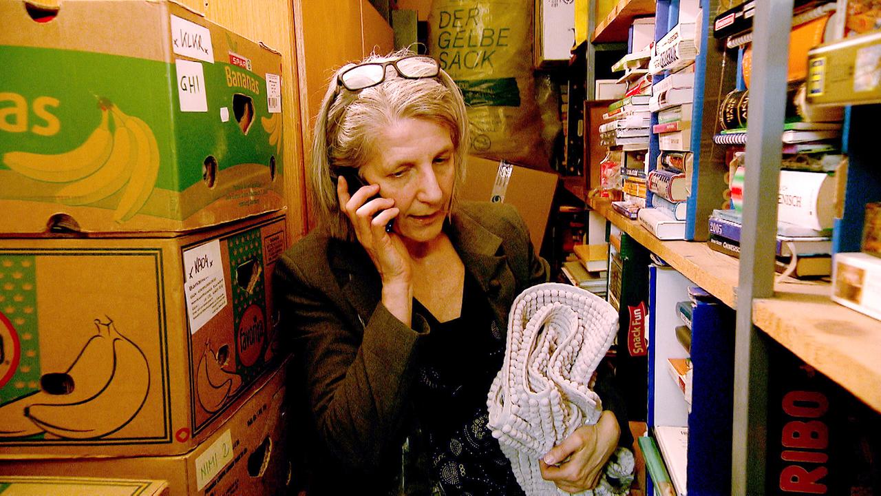 Am Bild ist Sabine, eine Frau mit Brille, die gerade in ihrem Keller telefoniert. In den Regalen sind viele Bücher. Auf der anderen Seite sind jede Menge Bananenkisten. 