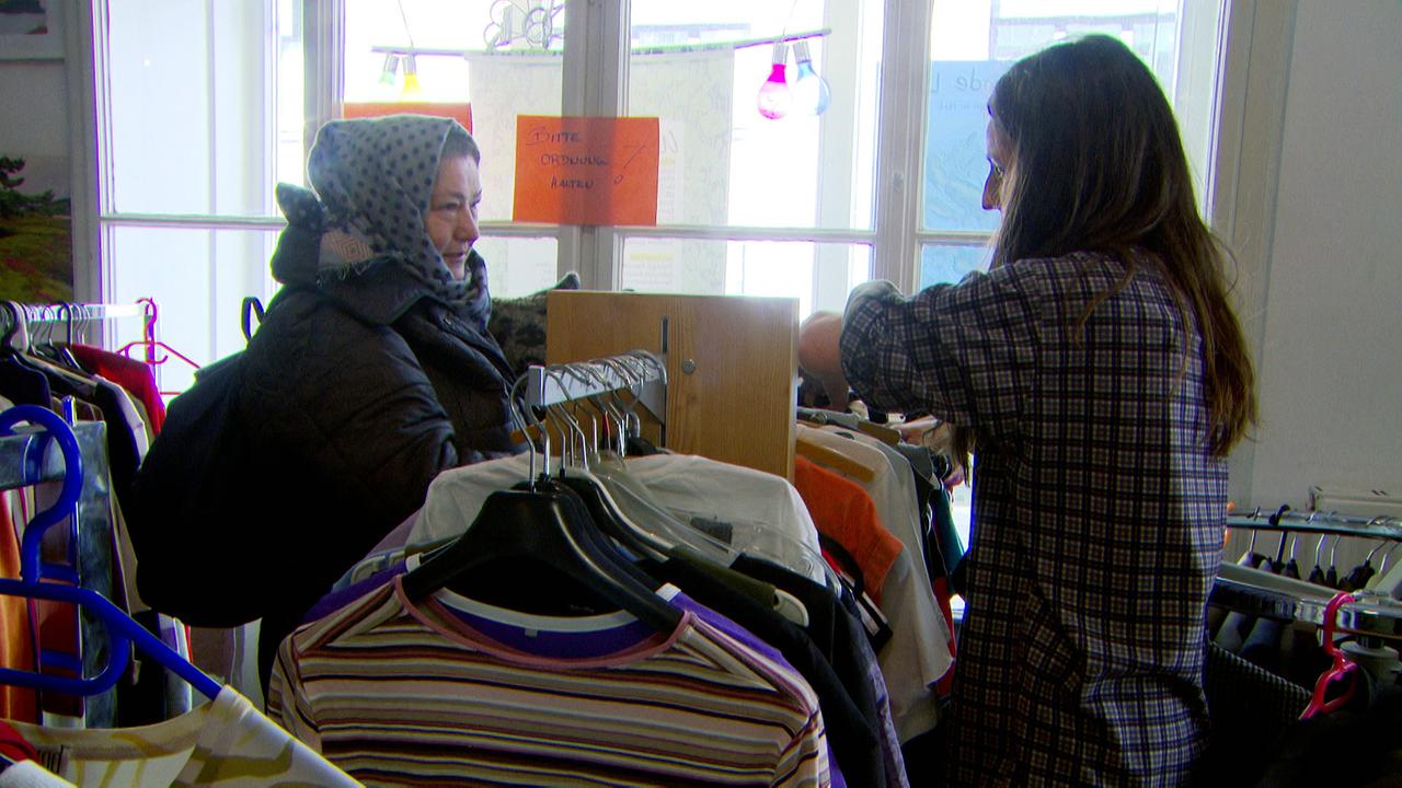 Am Bild sind zwei Frauen, die im Kostnix Laden in Salzburg zwischen einigen gut gefüllten Kleiderständern stehen.