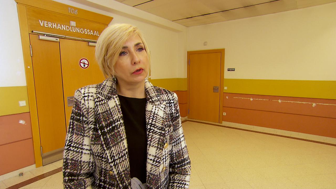© ORF Am Bild steht Frau Natalja vor dem Verhandlungssaal bei Gericht. Sie hat kurze blonde Haare, trägt einen karierten Blazer und ein schwarzes Oberteil.