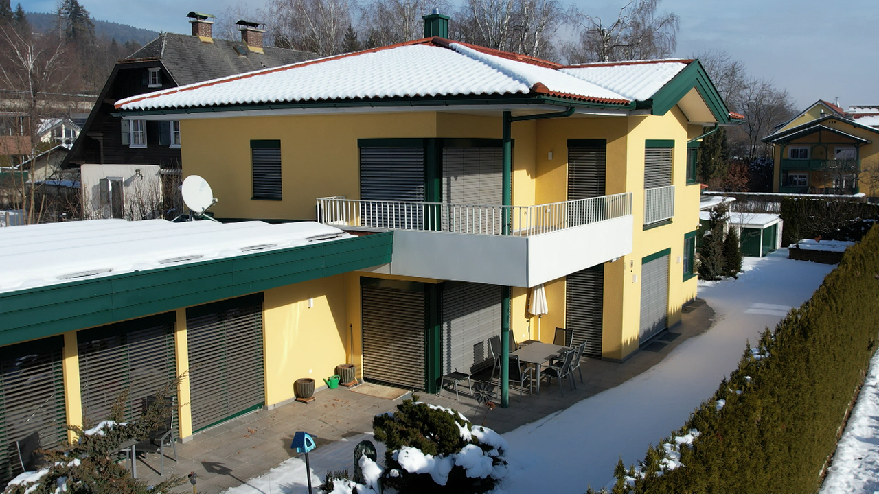 Am Bild ist die Villa in Velden. Ein großes, einstöckiges gelbes Haus mit grünen Säulen und einem großem Balkon.