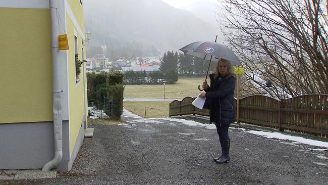 Frau H. steht mit einem Schirm am Zufahrtsweg zu ihrem Haus. Links ist das gelbe Haus der Nachbarn zu sehen.