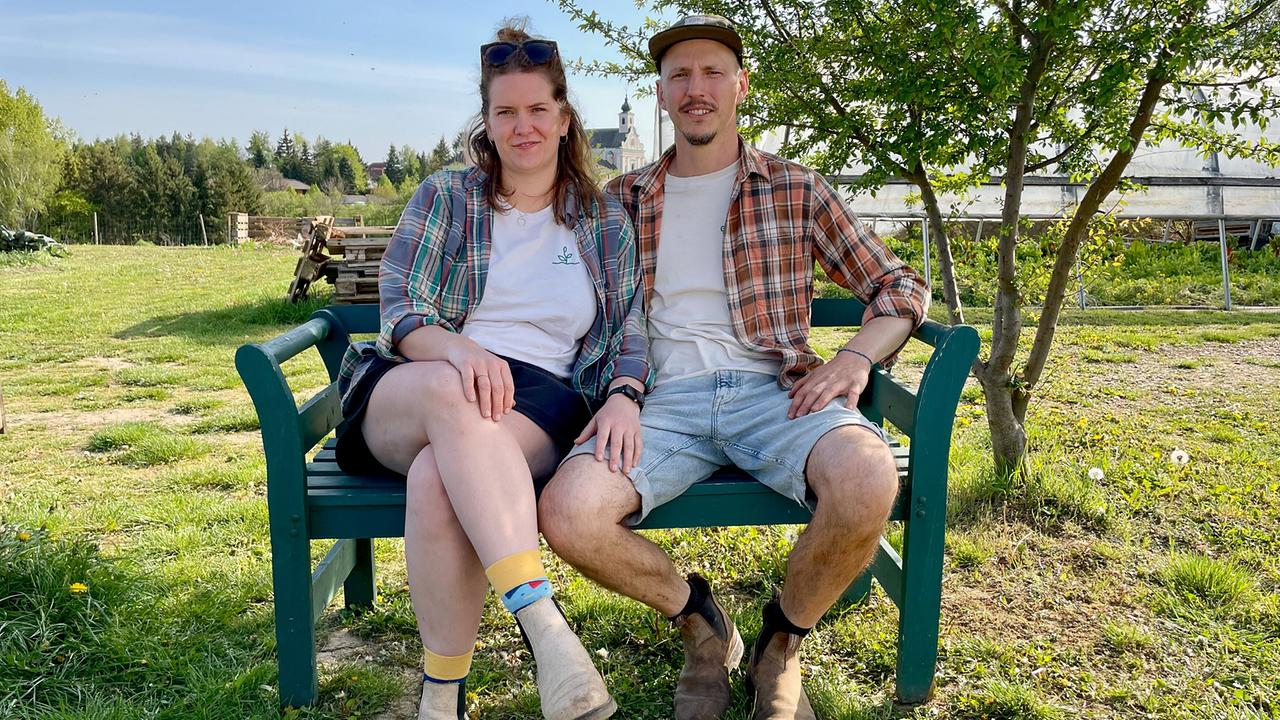 Die Gemüsebauern Verena und Michael Kietreiber sitzen in ihrem Gemüsegarten auf einer Bank vor Bäumen.