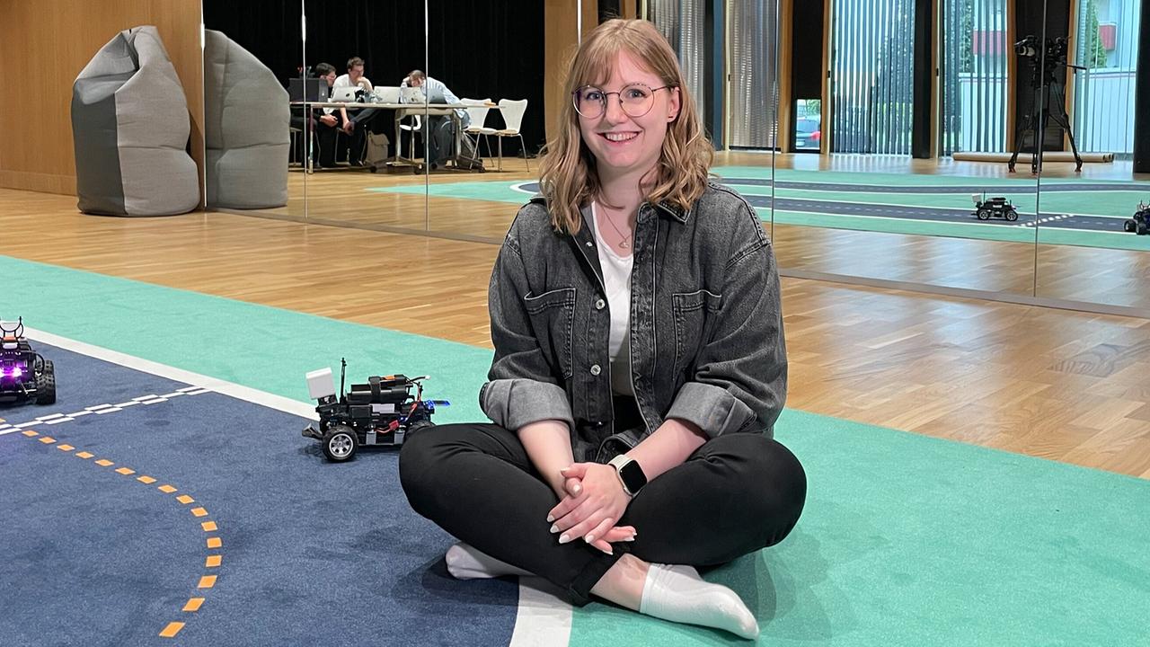 Studentin Alina Harant sitzt auf einem Teppichboden mit kleine Roboterfahrzeugen.