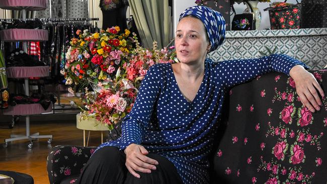 Susanne Bisovsky sitzt auf einem geblümten Sofa vor einem Hintergrund mit bunten Blumenarrangements und Modeobjekten.