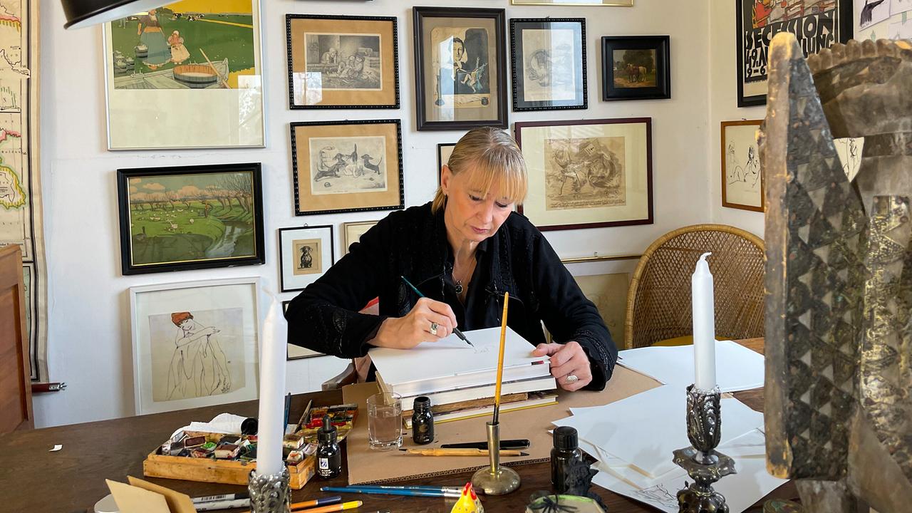 © ORF/Satel Film/David Haslinger Anette Smolka-Woldan sitzt an einem Tisch und zeichnet, umgeben von gerahmten Bildern an der Wand und verschiedenen Zeichenutensilien