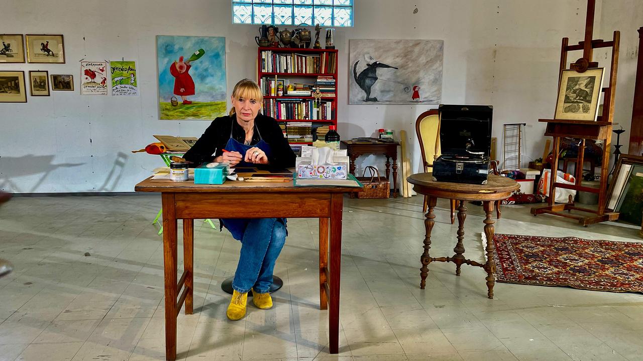 © ORF/Satel Film/David Haslinger Künstlerin Anette Smolka-Woldan sitzt an einem Holztisch in ihrem Atelier mit mehreren Gemälden im Hintergrund.