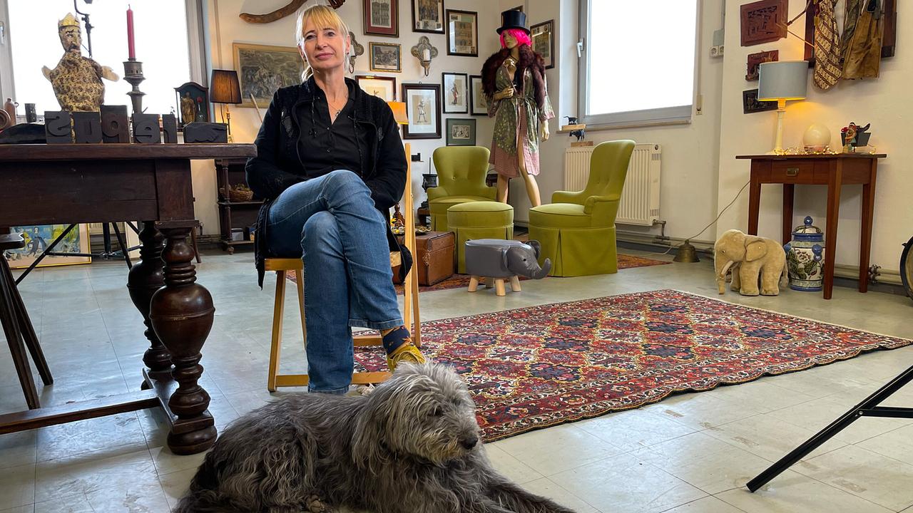 © ORF/Satel Film/David Haslinger Anette Smolka-Woldan sitzt mit ihrem Hund auf einem Teppich in ihrem Atelier mit Kunstwerken und Möbeln