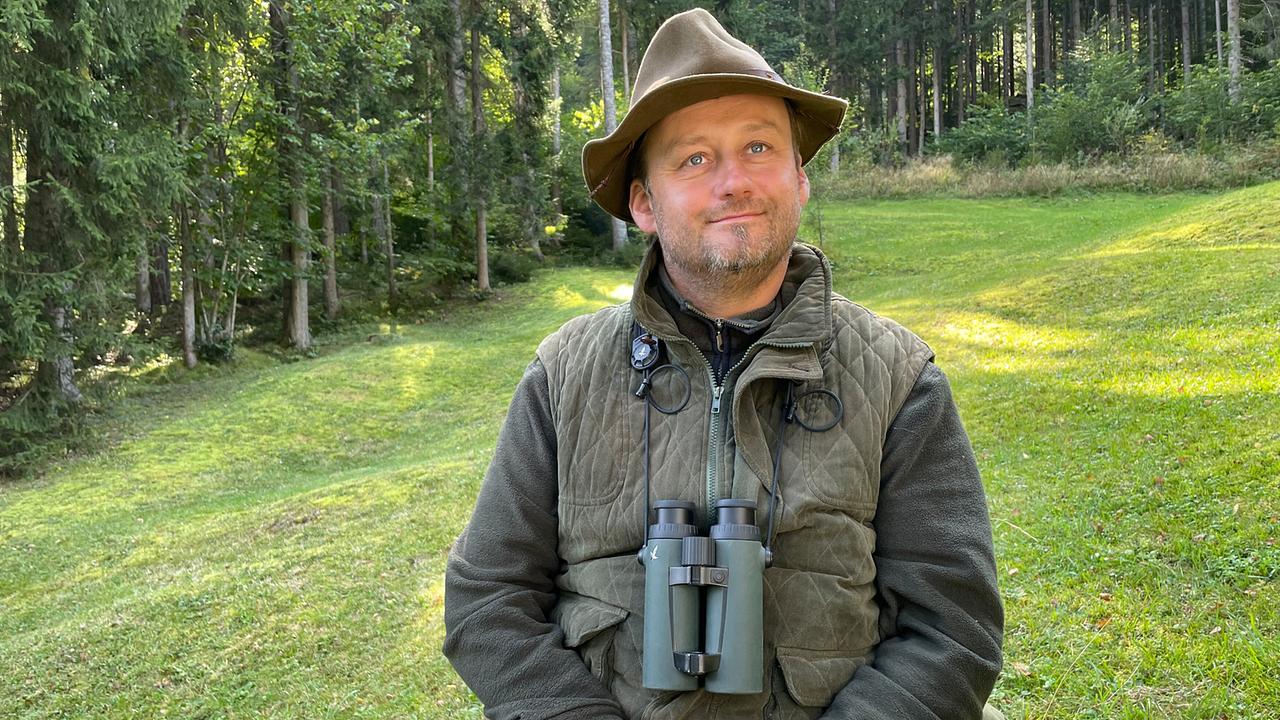 © ORF/Satel Film/David Haslinger Michael Winkler mit Hut und Fernglas um den Hals sitzt auf einer Wiese am Waldrand.