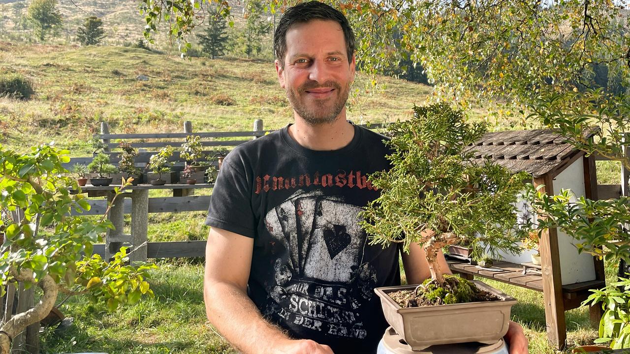 © ORF/Satel Film/David Haslinger Stefan Rindler steht im Freien und hält einen Bonsai-Baum in einem rechteckigen Topf vor sich.