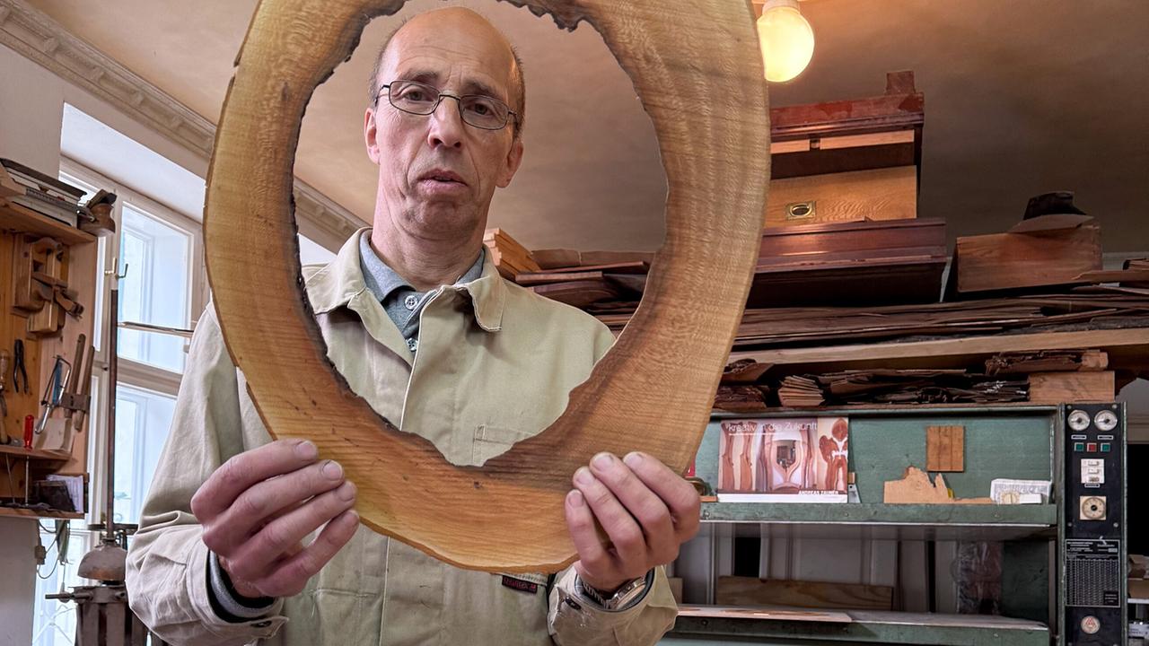 Andreas Zauner in seiner Werkstatt, er hält einen großen, runden Holzrahmen mit unregelmäßiger Maserung