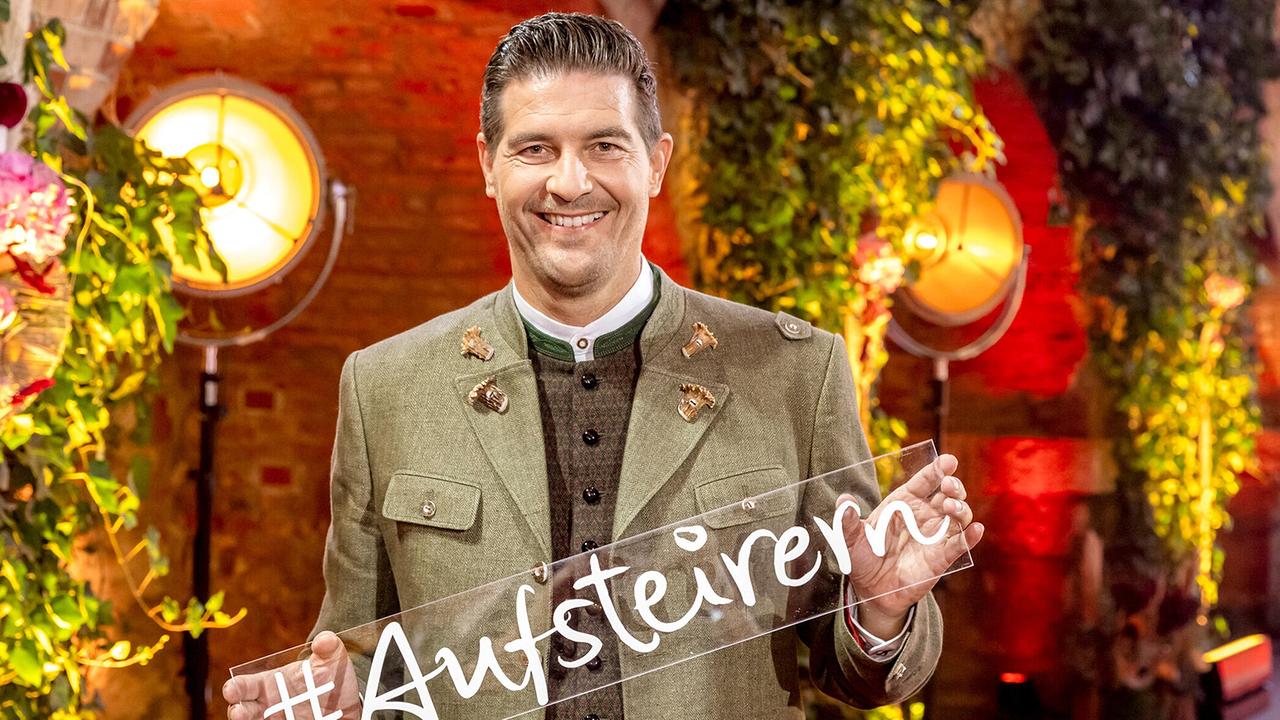 © ORF/Landessstudio Steiermark/Regine Schoettl Nobert Oberhauser in grüner Trachtenjacke hält vor einer üppig begrünten, warm ausgeleuchteten Kulisse ein Schild mit der Aufschrift „#Aufsteirern“ in die Kamera.