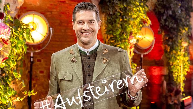 © ORF/Landessstudio Steiermark/Regine Schoettl Nobert Oberhauser in grüner Trachtenjacke hält vor einer üppig begrünten, warm ausgeleuchteten Kulisse ein Schild mit der Aufschrift „#Aufsteirern“ in die Kamera.