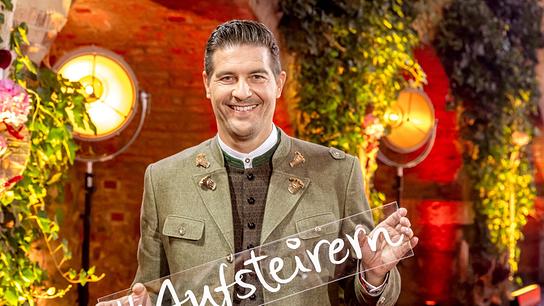 Nobert Oberhauser in grüner Trachtenjacke hält vor einer üppig begrünten, warm ausgeleuchteten Kulisse ein Schild mit der Aufschrift „#Aufsteirern“ in die Kamera.