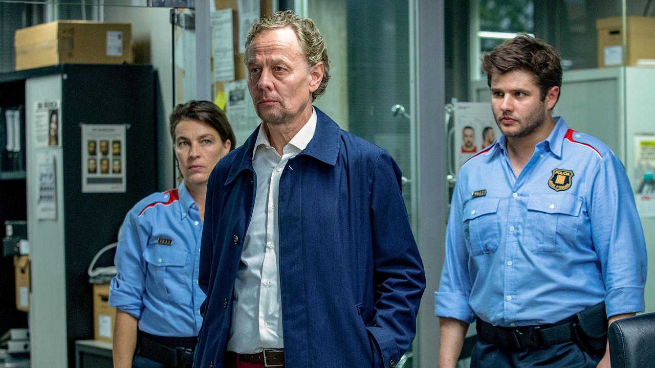 Ein Mann in blauem Mantel steht vor einer Frau und einem Mann in Polizeinuniform in einer Polizeiwache - er blickt ernst nach vorne