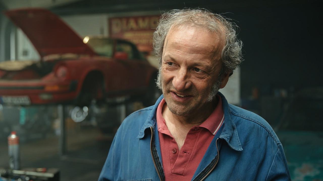 Max Schmid (Rolle: Mechaniker Detlef) im Interview in einer Werkstatt vor einem angehobenen roten Auto mit offener Motorhaube.