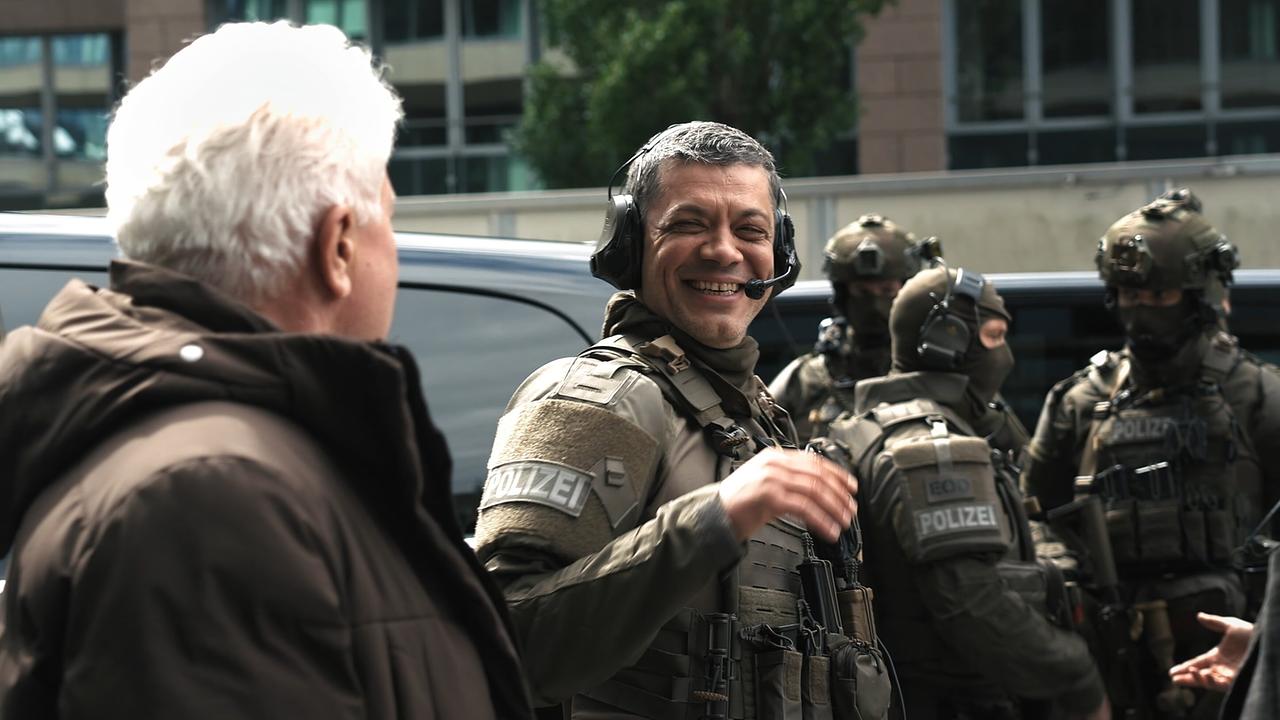 Carlo Ljubek (Rolle: Nikola Buvak) in schwerer Polizeiausrüstung  schaut am Set lachend zu Miroslav Nemec (Rolle: Ivo Batic), im Hintergrund weitere schwer ausgerüstete Polizeibeamte. 