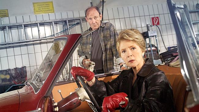  Christiane Hörbiger sitzt in ihrem Auto in einer Werkstatt, im Hintergrund Michael Mendl mit einem ölverschmierten Fetzen