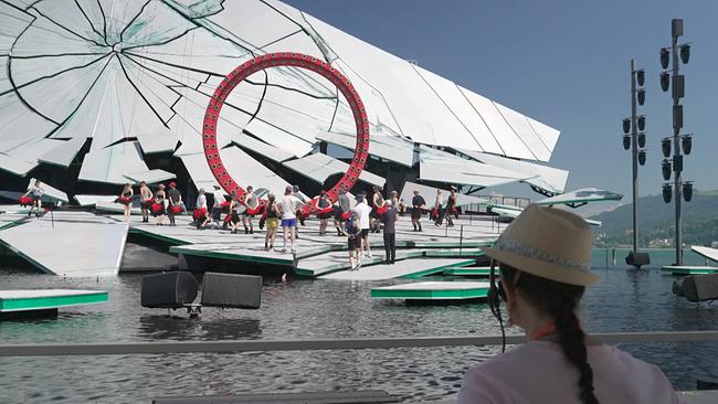 Auf einer schwimmenden Seebühne in Bregenz proben zahlreiche Mitwirkende vor einer monumentalen, zerbrochen wirkenden Kulisse mit rotem Ring.