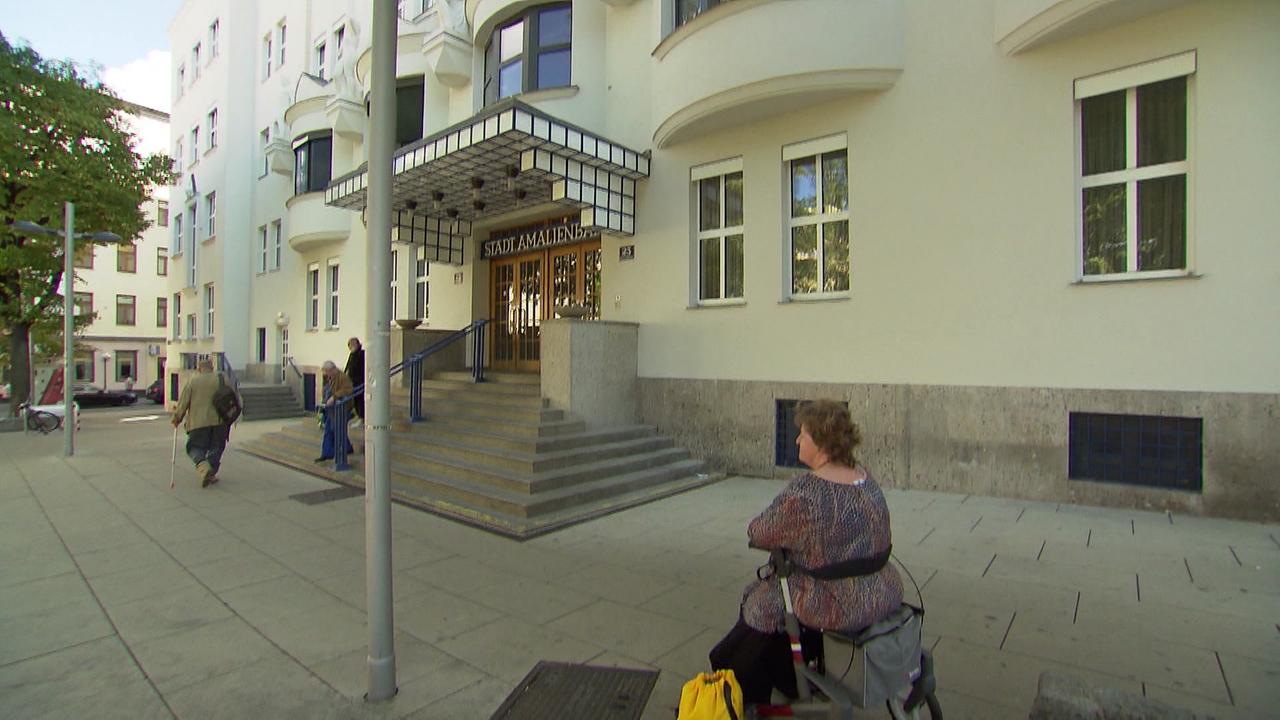 Auf dem Bild ist der Eingang zum Amalienbad (einem öffentlichen Schwimmbad in Wien). Eine Frau sitzt auf ihrem Rollator und wartet. Zum Eingang hin sind ungefähr 10 Stufen und keine Rampe.