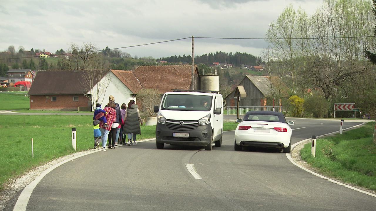 Auf einer Landstraße gehen am linken Rand eine Gruppe Eltern mit Kindern. Ein Bus und ein Auto fahren gerade vorbei. Man sieht, wie eng es wird, wenn Fußgänger und Autos einander begegnen. Bei erhöhter Geschwindigkeit der Fahrzeuge ist das eine Gefahr für die Fußgänger.