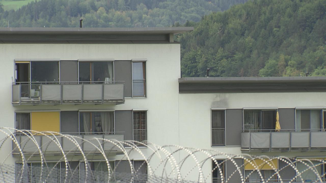 Im Vordergrund ist die Stacheldrahtumzäunung des Gefängnisses zu sehen, im Hintergrund ein Wohnhaus