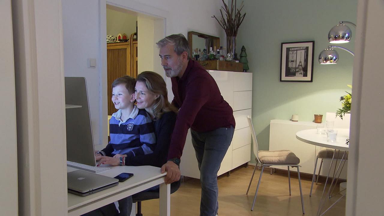 Ein Bub sitzt am Schoß seiner Mutter und sieht in den Computer. Der Vater steht hinter ihnen.