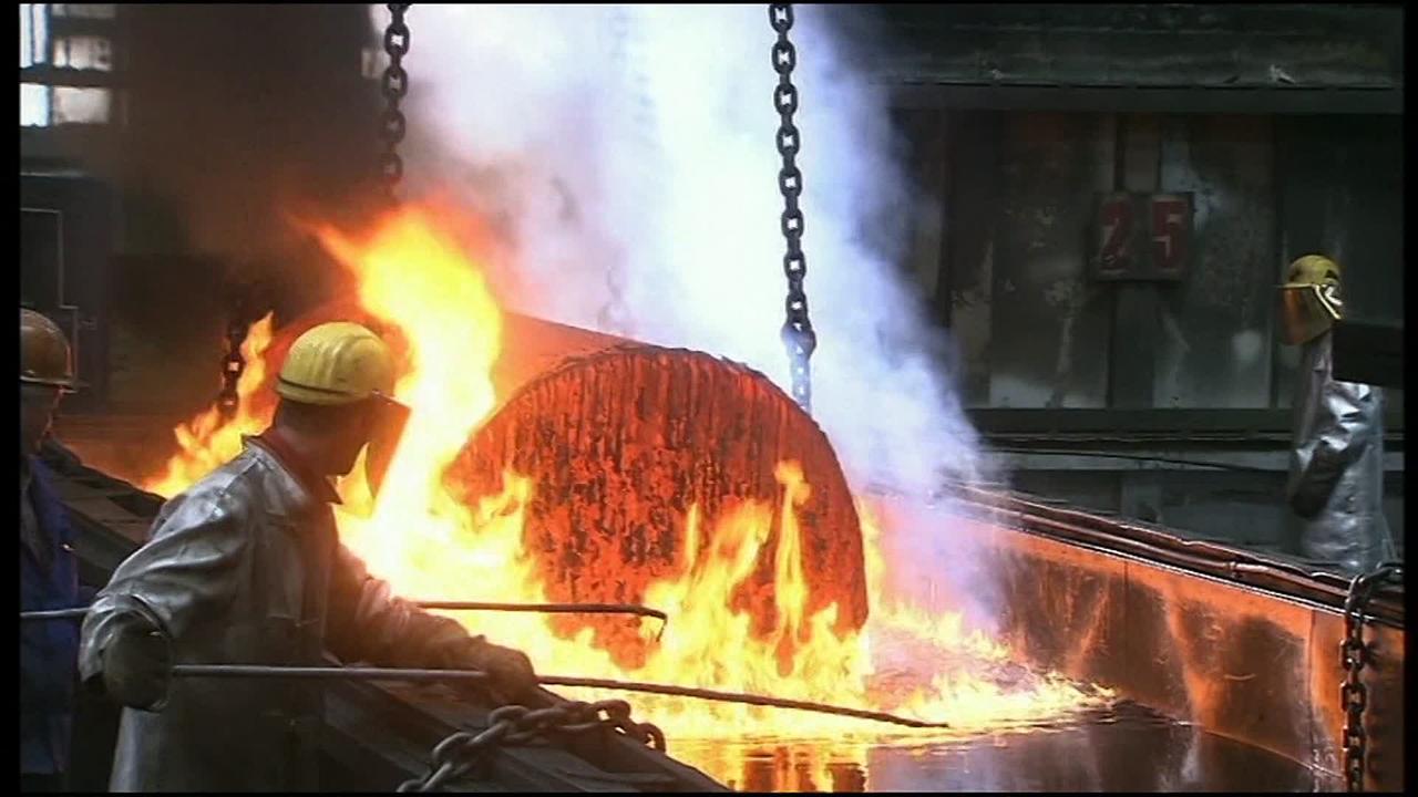 Arbeiter aus dem Voest-Werk bearbeiten ein großes brennendes Metallstück.